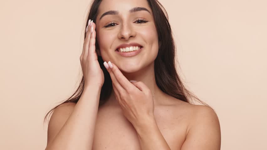 Close-up of a cheerful young Armenian woman with perfect skin, smiling and touching her face gently against a neutral background.