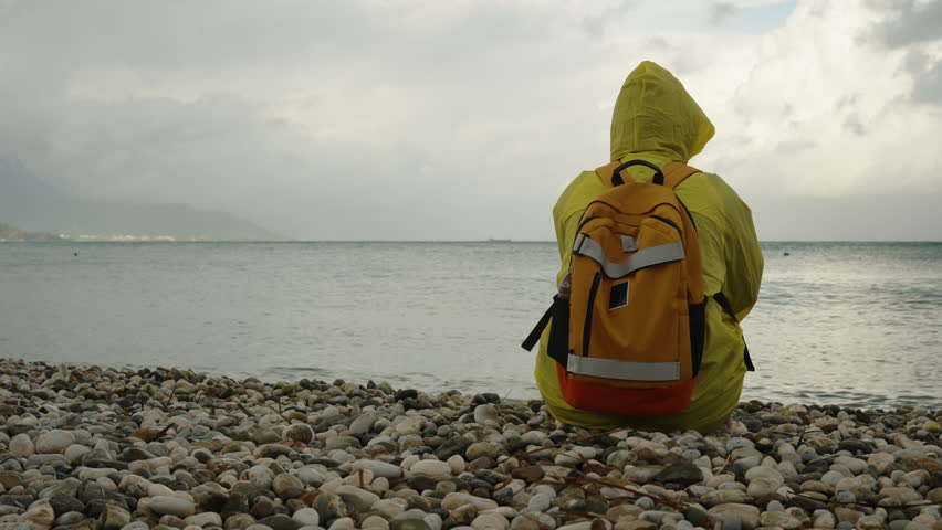 View from behind of a person in a yellow raincoat with an orange backpack, sitting on the beach by the stormy sea. Traveler.
