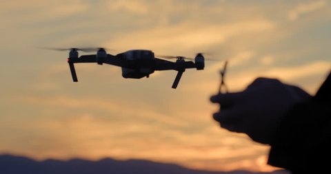 Close view of drone pilot's hands as he controls quadcopter hovering then flying off in the background. Aircraft and pilot silhouetted against sunset sky. Slow motion recorded at 60fps, 4K.