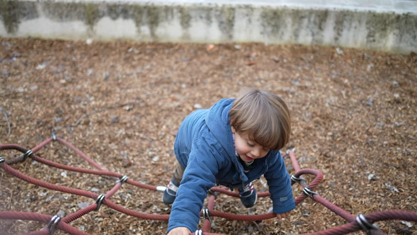 Happy active child climbing playground ropes during fall season. Autumn day play of 3 year old boy having fun outdoors, wearing blue jacket showing physical effort