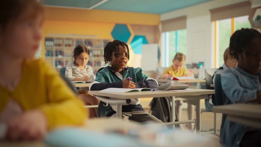 Portrait of a Cute Little African Boy with Stylish Hair Sitting Behind a Desk in Class in Elementary School. Young Pupil is Focused on a Lecture, Listening to a Teacher with Other Kids Royalty-Free Stock Footage #3419488243