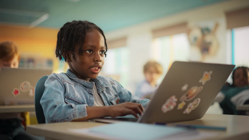 Portrait of a Cute Little African Boy with Stylish Hair Sitting Behind a Desk in Class in Elementary School. Young Pupil Using a Laptop Computer while Listening to a Teacher with Other Kids - Powered by Shutterstock - Get 15% off with code: PIKWIZARD15
