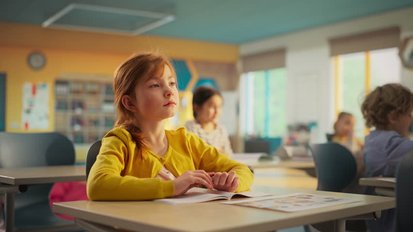 Portrait of a Cute Smiling Redhead Girl Sitting Behind a Desk in Class in Elementary School. Young Pupil is Looking at Camera, Smiling. Kids Educated in Modern Primary School