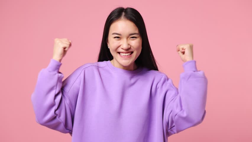 Excited jubilant overjoyed happy young woman of Asian ethnicity wear purple sweatshirt do winner gesture celebrate clench fists say yes isolated on plain pastel light pink background studio portrait