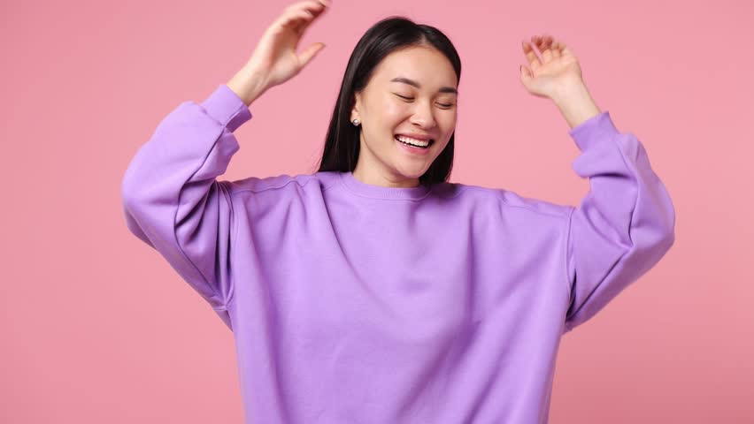 Fancy vivid happy bright young woman of Asian ethnicity wear purple sweatshirt dance waving rising have fun enjoy relax celebrate play isolated on plain pastel light pink background. Lifestyle concept