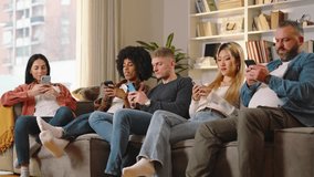 Group of multi-ethnic friends, all smiles, chat and laugh on a sofa during a lively video call. Greetings are exchanged with waves, showcasing their strong bond even through the digital connection