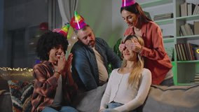 Friends Surprise Birthday Girl Bringing Cake At Indoor House Party. Face Of Overjoyed Woman Lights Up With Joy As She Eagerly Blows Out The Burning Candle In A Moment Of Celebration. 