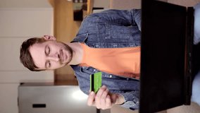 Portrait of smiling young man holding debit credit card in hand and using laptop in living room. Vertical video.