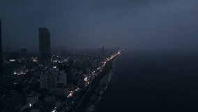 Aerial night view of Colombo cityscape with illuminated streets, urban architecture alongside coast. Drone footage reveals ocean waves, dynamic skyline, and traffic in Sri Lanka capital at dusk.