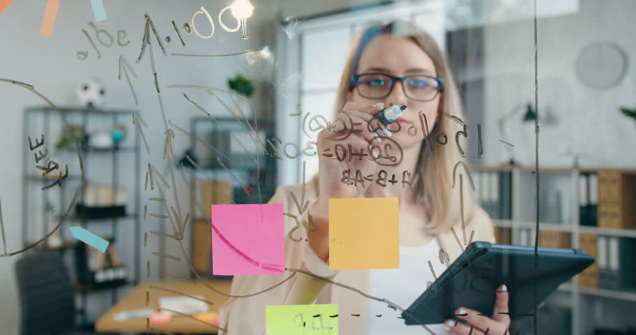 Attractive woman in glasses writing mathematical calculations on glass board in modern office,creativity concept