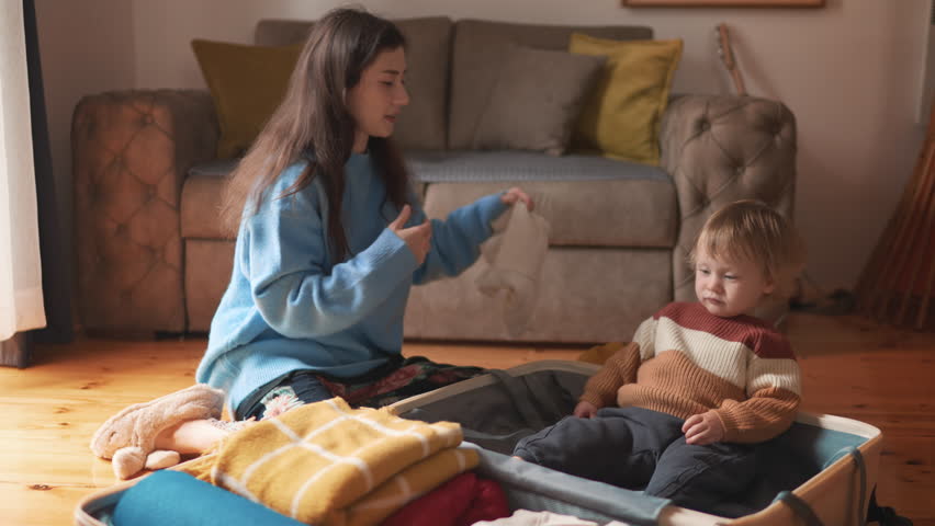 Asian mother and her toddler son preparing for their travel adventure, packing clothes into suitcase, getting ready for road trip travelling, happy family concept. Mom and a kid pack luggage together.