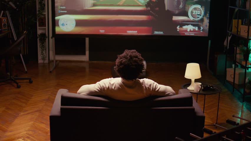 Close up shot of african american man watching esports competition on gigantic smart TV, relaxing on couch. Gamer in home theatre following gaming tournament on television set
