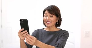 Happy attractive woman smiling enjoying face time video chat talking using mobile phone relaxing standing at home. Mature asian beauty streaming internet blogger for communication palm background.