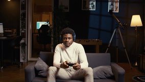 African american man playing shooter videogame in warm apartment, holding controller. Gamer participating in PvP online multiplayer game using console system and headphones for immersive experience