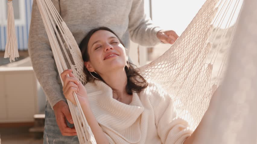 A young woman relaxes in a hammock, closing her eyes in pleasure as her partner embraces her from behind, sharing a warm, affectionate moment.
