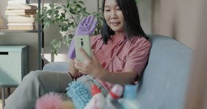 Cheerful Asian woman wearing rubber gloves making online video call speaking waving hand using smartphone after cleaning house. Communication and domestic chores concept.