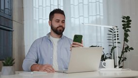 Caucasian man having video call by smartphone in green case and waving good bye while sitting at domestic workplace. Remote worker having break from researching and talking to family on mobile gadget.
