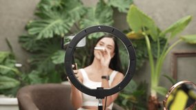 Video zoom in through center of circular ring light revealing woman use selfie stick for beauty and cosmetic tutorial video content. Beauty blogger amidst serene ambiance of natural garden. Blithe