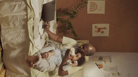 Vertical side full footage of African American parent sitting on bed and working on laptop while bottle feeding little two-tear-old Biracial daughter in decorated bedroom in evening