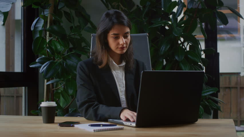 A young business woman makes a call on her smartphone in a modern office with plants. A female manager holds a phone in her hands behind a laptop in a coworking space. A woman is having talk with boss