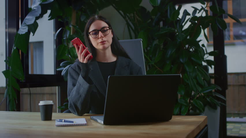 A young business woman makes a call on her smartphone in a modern office with plants. A female manager holds a phone in her hands behind a laptop in a coworking space. A woman is having talk with boss Royalty-Free Stock Footage #3420181053