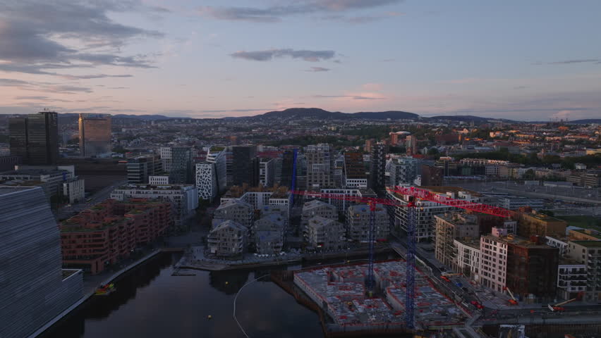 Forwards fly above modern multistorey buildings in urban borough against color twilight sky. Tower cranes on construction site. Oslo, Norway