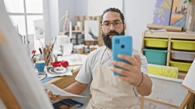 Bearded man in studio holding artwork and smartphone capturing selfie