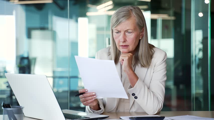 Focused elderly senior gray haired businesswoman in a suit reads a document sitting at a workplace in a business office. Thoughtful mature female financier is busy with paper work, looks at the report Royalty-Free Stock Footage #3420273489