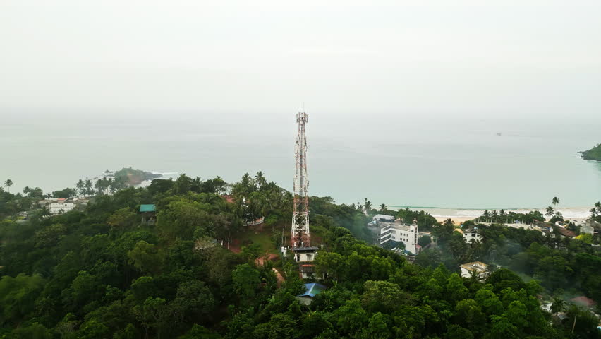 Aerial view captures towering communications mast rising above lush island foliage, overlooking coastal village, beach, and serene sea to ensure connectivity.