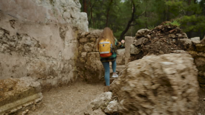 Ruins of ancient houses in the city of Phaselis, remnants of stone walls in the forest through which a young woman with a backpack is walking.