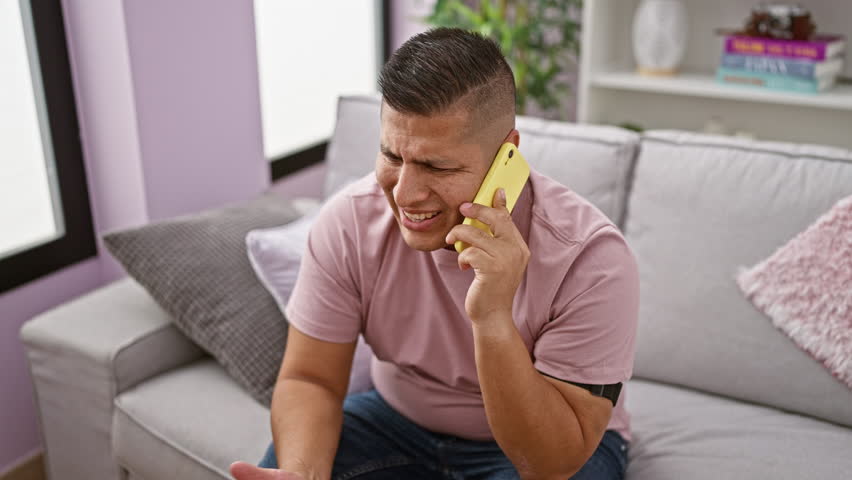 Worried young latin man caught in serious disagreement at home, angrily venting on smartphone while sitting on the living room sofa, indoor background Royalty-Free Stock Footage #3420458853