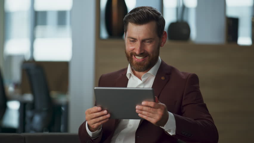 Smiling manager looking tablet computer screen in business office interior cabinet. Cheerful bearded professional man swiping pages modern pad in big hall. Digital corporate marketing work concept.
