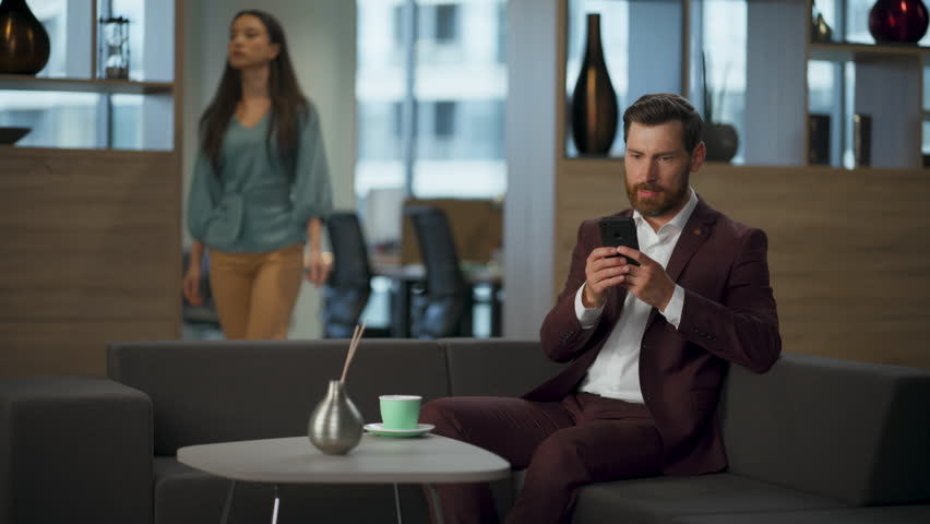 Annoyed business manager man typing message mobile phone office interior hall. Angry financial professional man moving hand looking screen smartphone big comfortable sofa. Stressful work concept.
