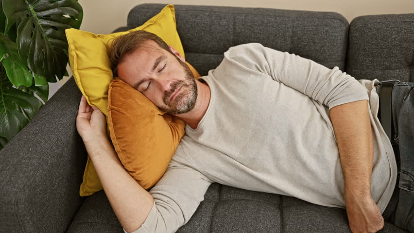 Middle-aged man napping on a couch with a yellow pillow and white blanket in a cozy home interior.