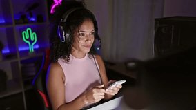 Stunning young hispanic female gamer streaming, speaking on smartphone while rocking headphones in a gaming room during a vibrant, virtual game night