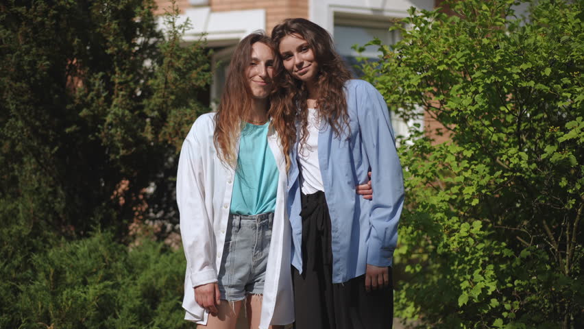 Two girls stand hugging in a city park on a bright sunny summer day among green trees. The girls smile and look at the camera