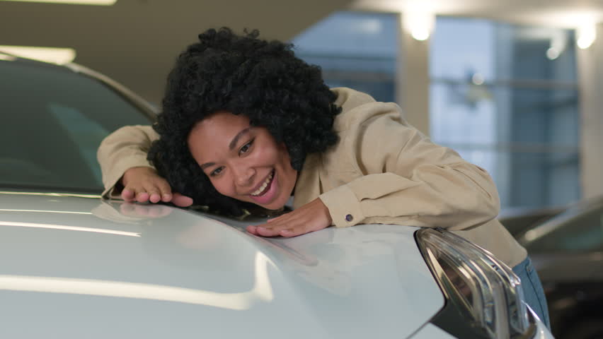 Happy woman African American lady female buyer client hugging hood of new modern car joyful smiling girl dreaming buying vehicle in auto showroom leaning on luxury automobile in store salon dealership