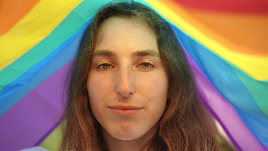 Close-up. The face of a girl who hid from the sun under a rainbow flag. The girl smiles and looks at the camera