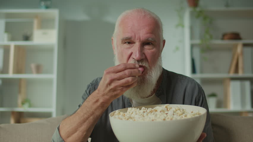 An Old Man Eating Popcorn and Concentrating on Watching TV.A Serious Elderly Man Sitting on the Sofa and Watching a Very Interesting Movie.Films as a Way of Emotional Outlet and Self-expression. Royalty-Free Stock Footage #3420639451