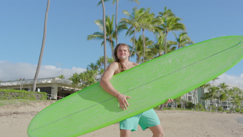 A joyful surfer with long hair smiles and fake guitar playing on a vibrant green surfboard on a sunny beach, with palm trees and a clear sky enhancing the tropical mood. Slow Motion, Camera 4K RAW.  Royalty-Free Stock Footage #3420796875