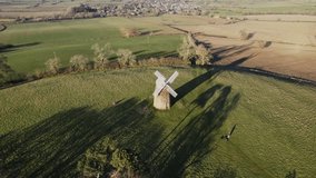 A 4K video of a slow arial orbit of a Stone Windmill with white wooden sails on a green grassy hill in rural England showing the hill and surrounding green countryside in Upper Tysoe in Warwickshire