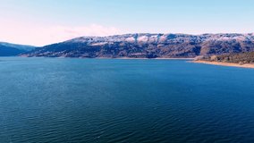 A drone captures the tranquil beauty of a mountainous winter landscape, with the calm waters of an adjacent lake reflecting the clear blue sky. national park Mavrovo in Macedonia