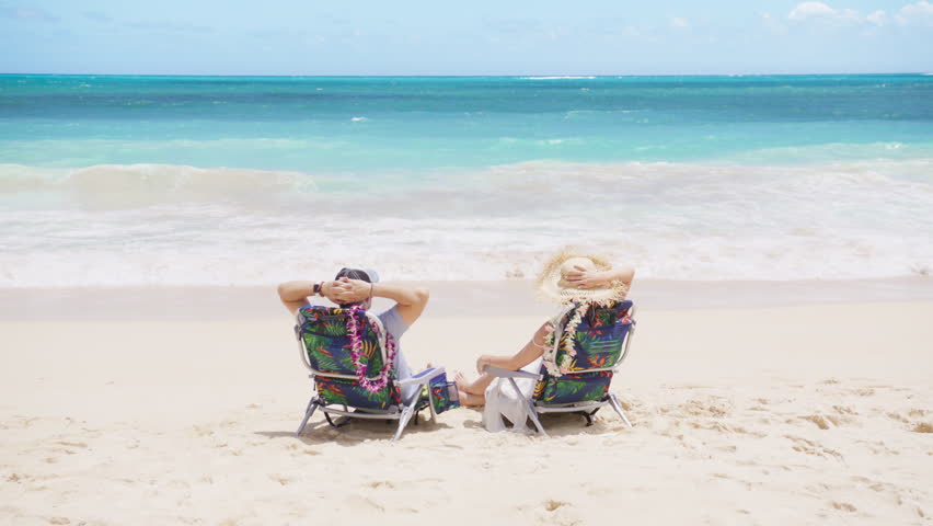 Back view of man and woman enjoy light blue ocean views and sun bathing together at summer holiday. Male and female relaxing outdoor lifestyle, sitting in beach chairs on tropical travel vacation Oahu