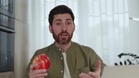 Handsome male blogger biting juicy red apple and enjoying it while sitting at table with wireless laptop at home. Emotional caucasian male agitating healthy lifestyles and natural products indoors.