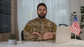 Confident army officer talking to camera while sitting with crossed arms by desk with portable computer American flag. Bearded caucasian male in camouflage giving motivational speech for soldiers.