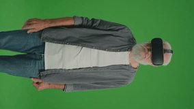 Vertical View.Green Screen. An Old Man With Gray Beard in VR Glasses Examines and Touches Everything Around. VR for the Socialization of the Elderly.