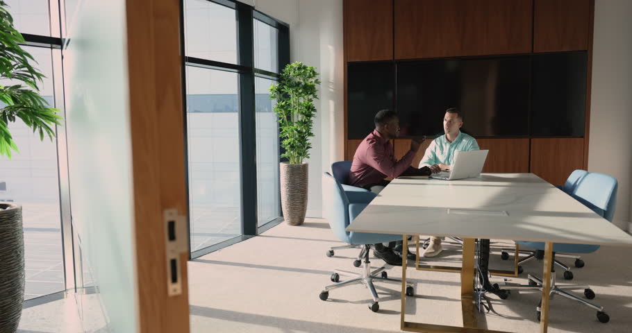 Two young multi-ethnic teammates talking met in modern conference room, coworkers engaged in teamwork, coach teaching intern, having business conversation with workmate, manager sell company services