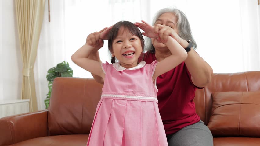 happy time Grandma and cute little Asian granddaughter in the living room at home. Granddaughter dancing for fun. family concept