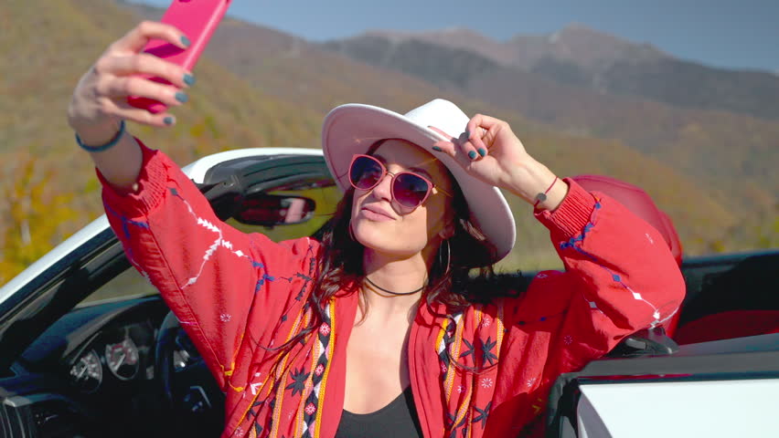 Succesful brunette woman wearing in boho style is making selfie on phone in mountains near the car. She is looking at smartphone camera and smiling. Tourism, journey, vacation, travel, car trip.