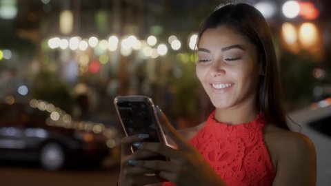 A young beautiful latina woman uses her phone to text and chat while she smiles. Busy street, cars, background lights at night. Concept apps for young adults friendship technology communication